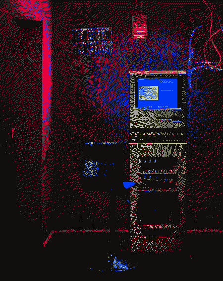 A dark room lit with neon red light and the blue light of a computer screen in the center, with a doorless exit at the left. The computer is very old-looking and tall, almost like a big plastic brick the height of a person. It has a small square screen at the top, a keyboard and some mechanical pieces below. There is a tall chair in front of the computer. There are some cables and a small poster that can't be seen very well in the wall.