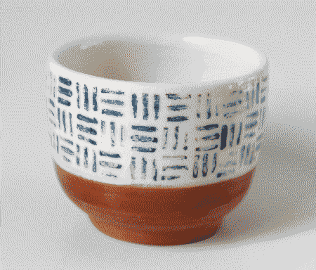 A small wheel-thrown cup. It is reddish in the underside, the natural color of the non-glazed terracotta. It is white from middle to top and inside, with stamped dark blue line motifs over the white. It is glazed over the white part.