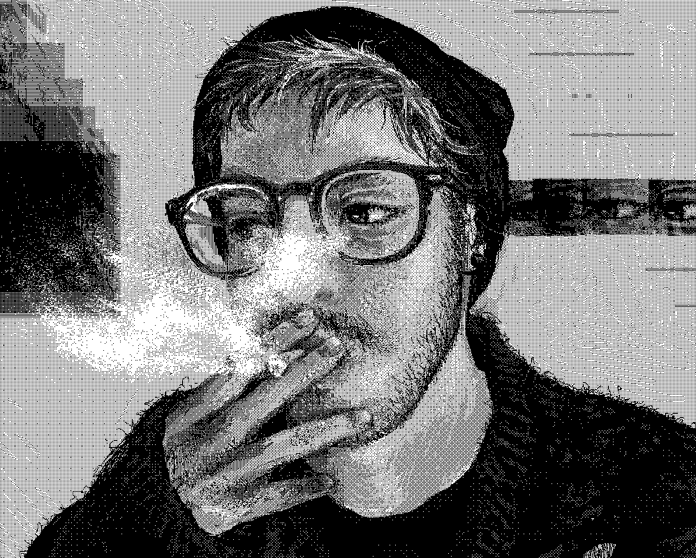 A digital black-and-white pixelled bust-up portrait of a young white man with glasses, short hair, a black beanie and a black yarn sweater. He has a moustache and stubble. He looks to his right side as he holds a cigarette in his mouth. Smoke is coming out of it, partially covering his face. His left eye is glitched and repeated horizontally many times to his left, out of his face and looking at him.